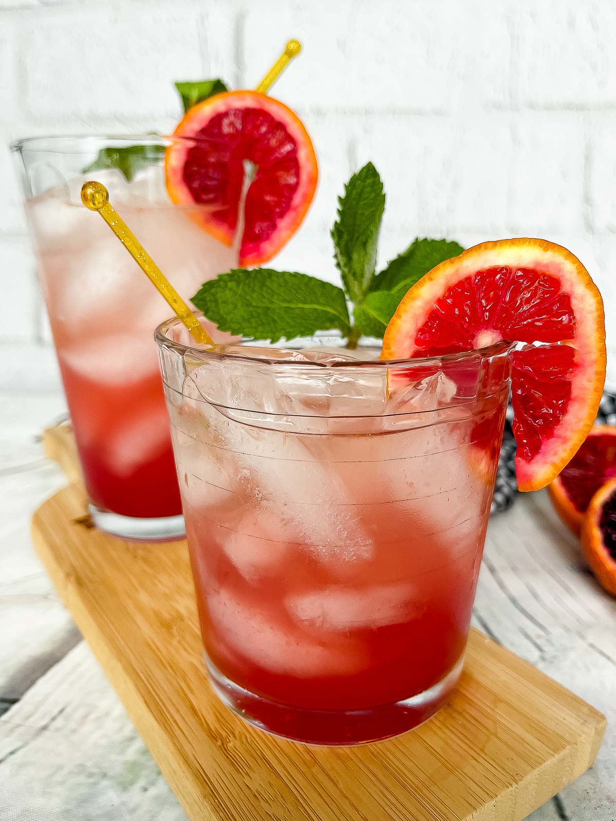 Two blood orange ginger cocktails in glasses with wedges of blood orange and mint sprigs on top of a wooden board with a garlic bulb on the side