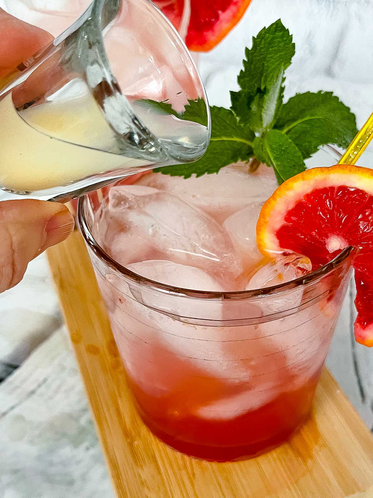 A blood orange ginger cocktail in a clear glass with a wedge of blood orange and mint sprig on top of a wooden board