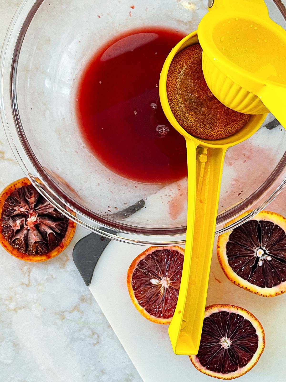 A yellow juice squeezer squeezing a blood orange into a clear glass bowl with orange halves on top of a cutting board on the side