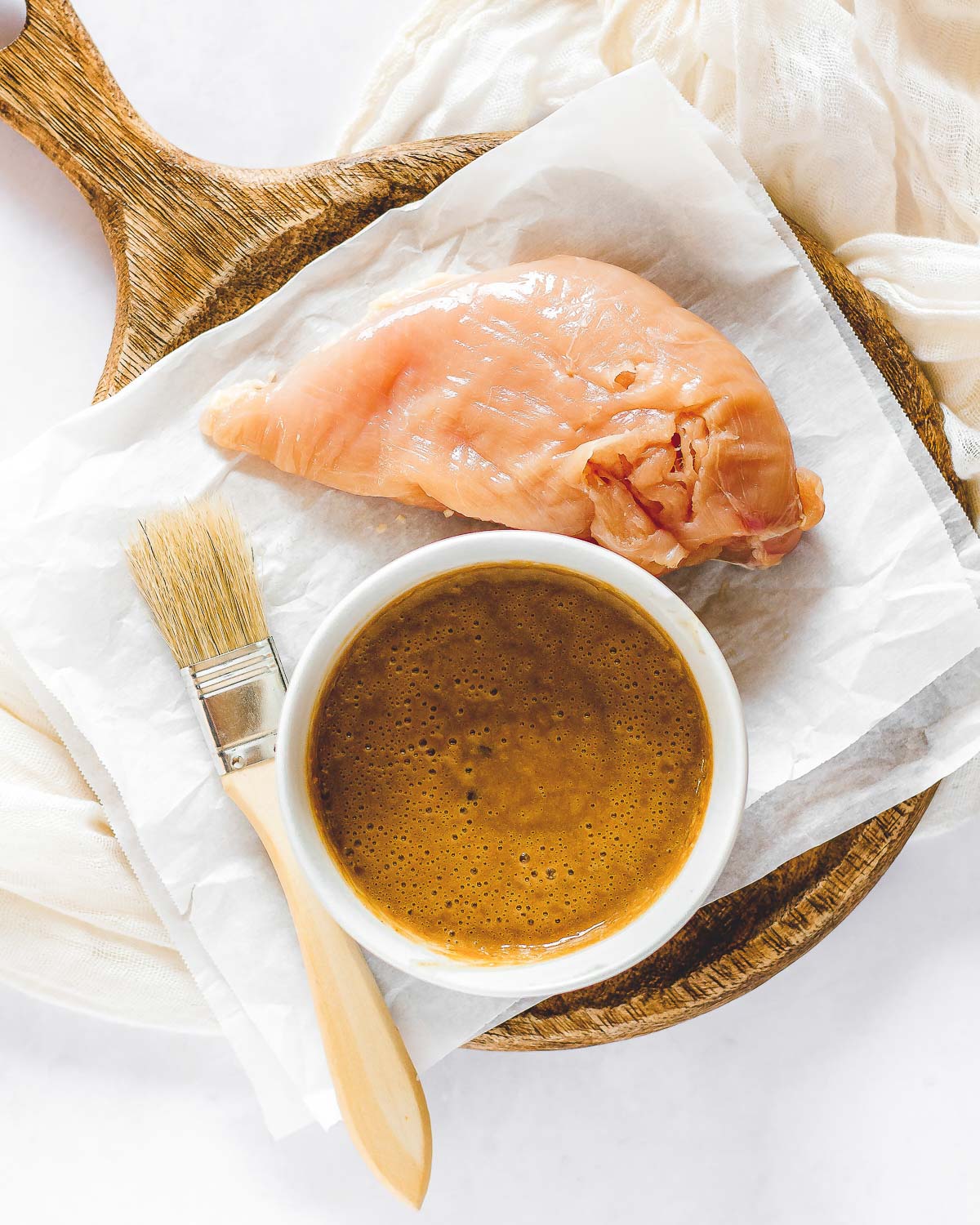 A chicken breast on wax paper next to a bowl of peanut sauce with a basting brush on the side on top of a wooden tray.