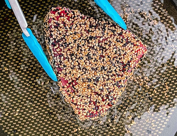 A tuna steak coated with sesame seeds searing in oil in a frying pan with a set of blue tongs.