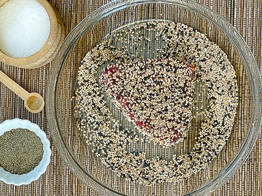 A raw tuna steak dredged with sesame seeds in a glass pie plate with salt and pepper on the side.