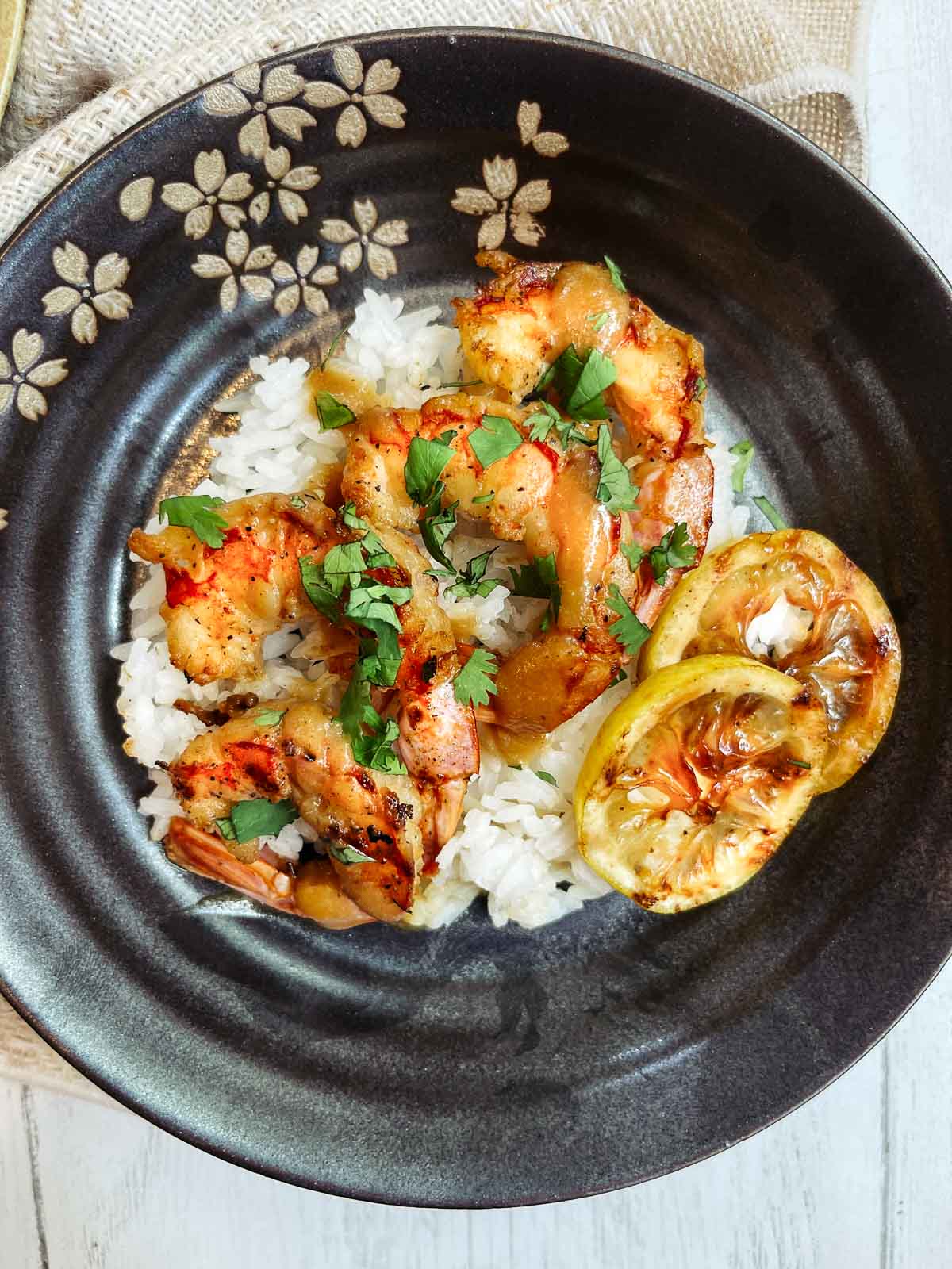 A black plate of grilled shrimp on top of white rice with charred lemon slices on top of a white wooden board.