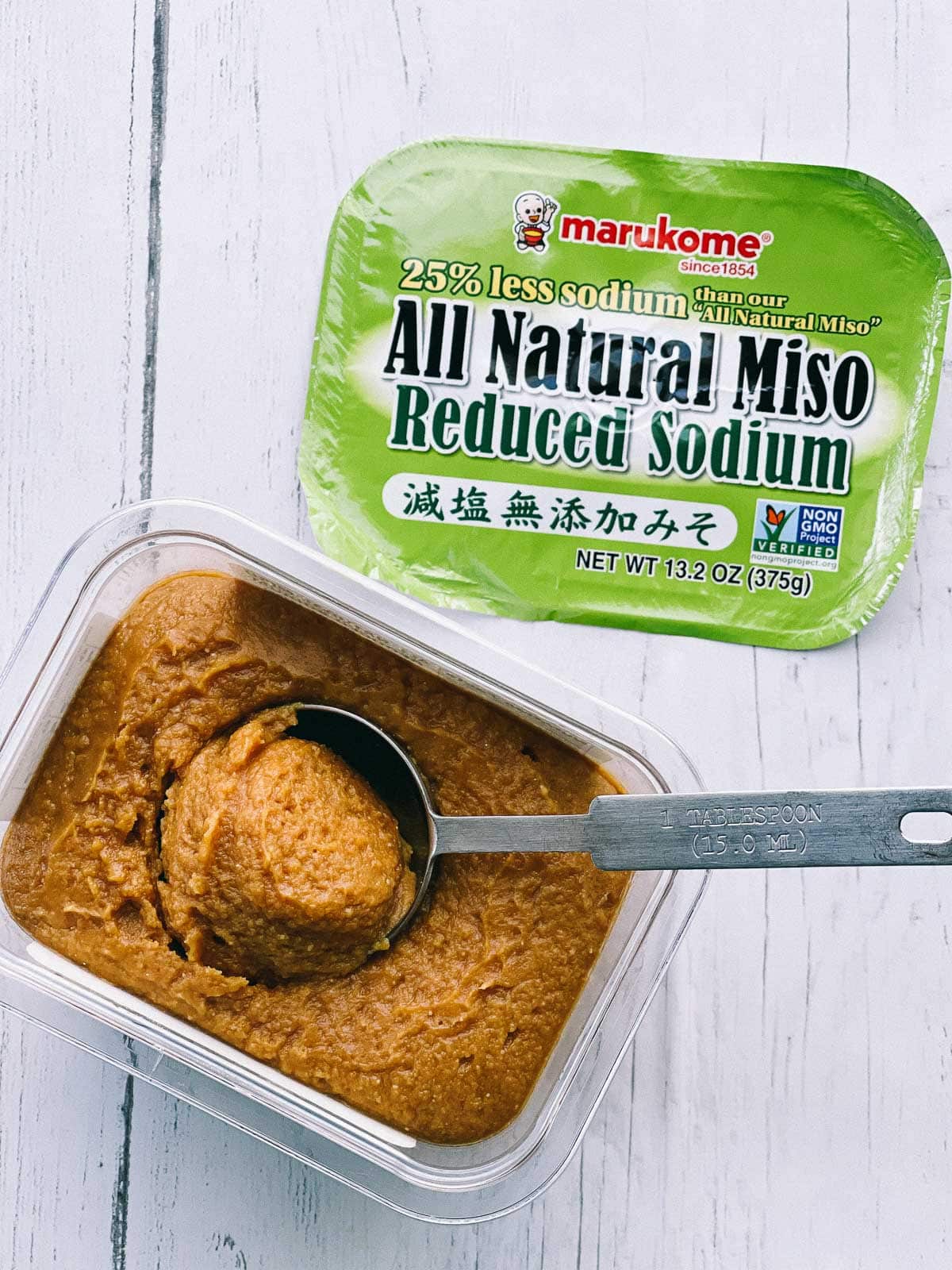 A container of miso paste with a measuring spoon inserted and the lid on the side on top of a white wooden board.