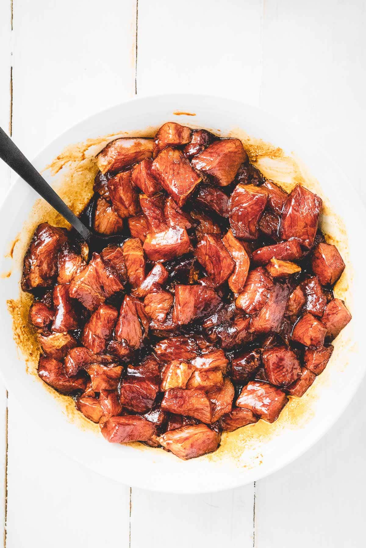 Pork chunks marinating inside a white bowl with a spoon on top of a white board.