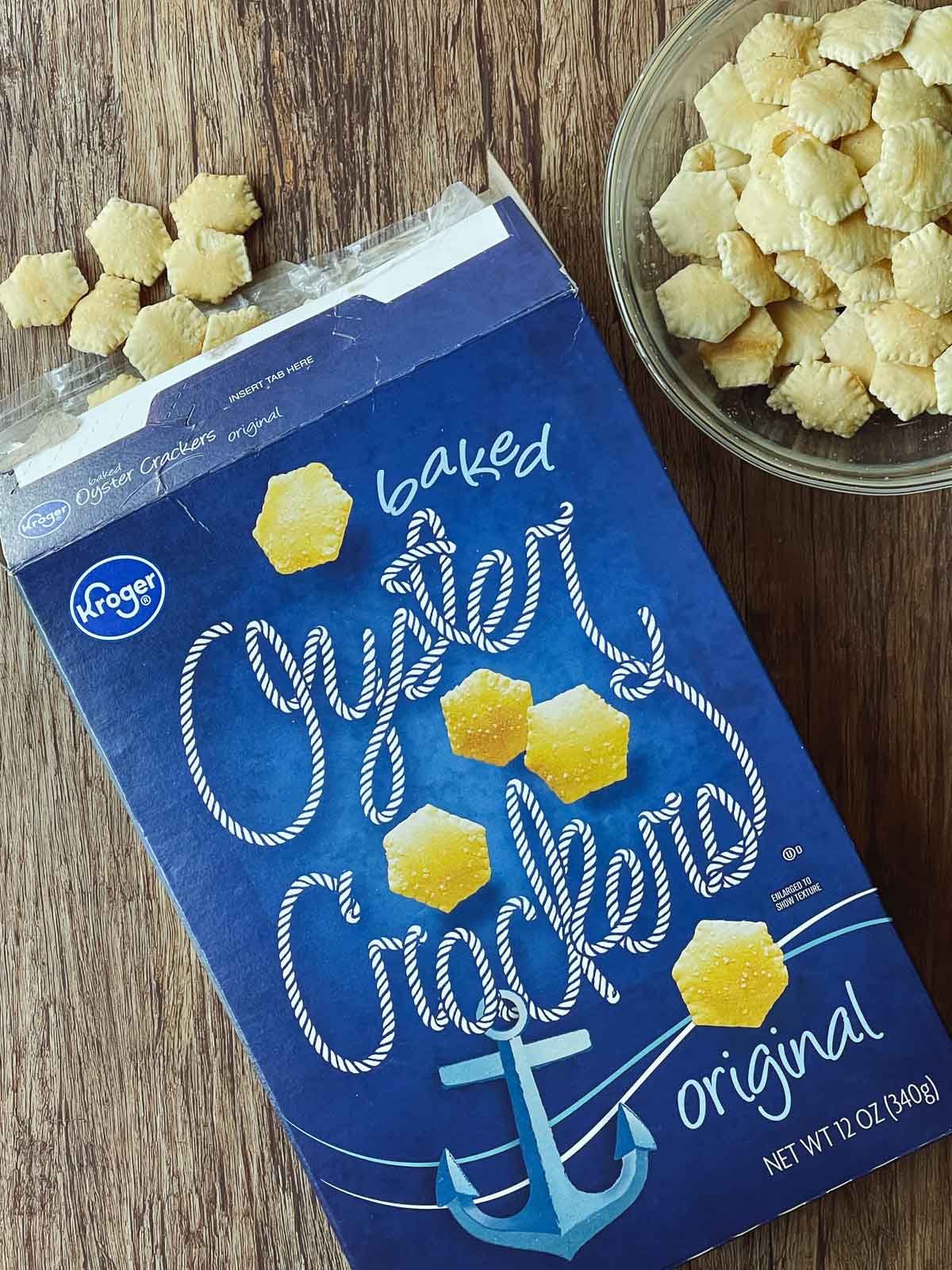 A blue box of oyster crackers on top of a wooden board with a glass bowl of oyster crackers on the side.