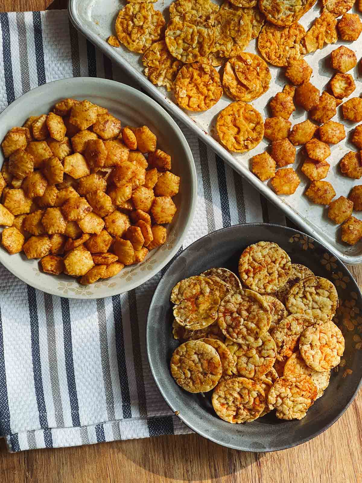 Sriracha popcorn cakes in a gray bowl and Sriracha oyster crackers in a light gray bowl on top of a striped kitchen towel and a baking tray with more crackers on the side.