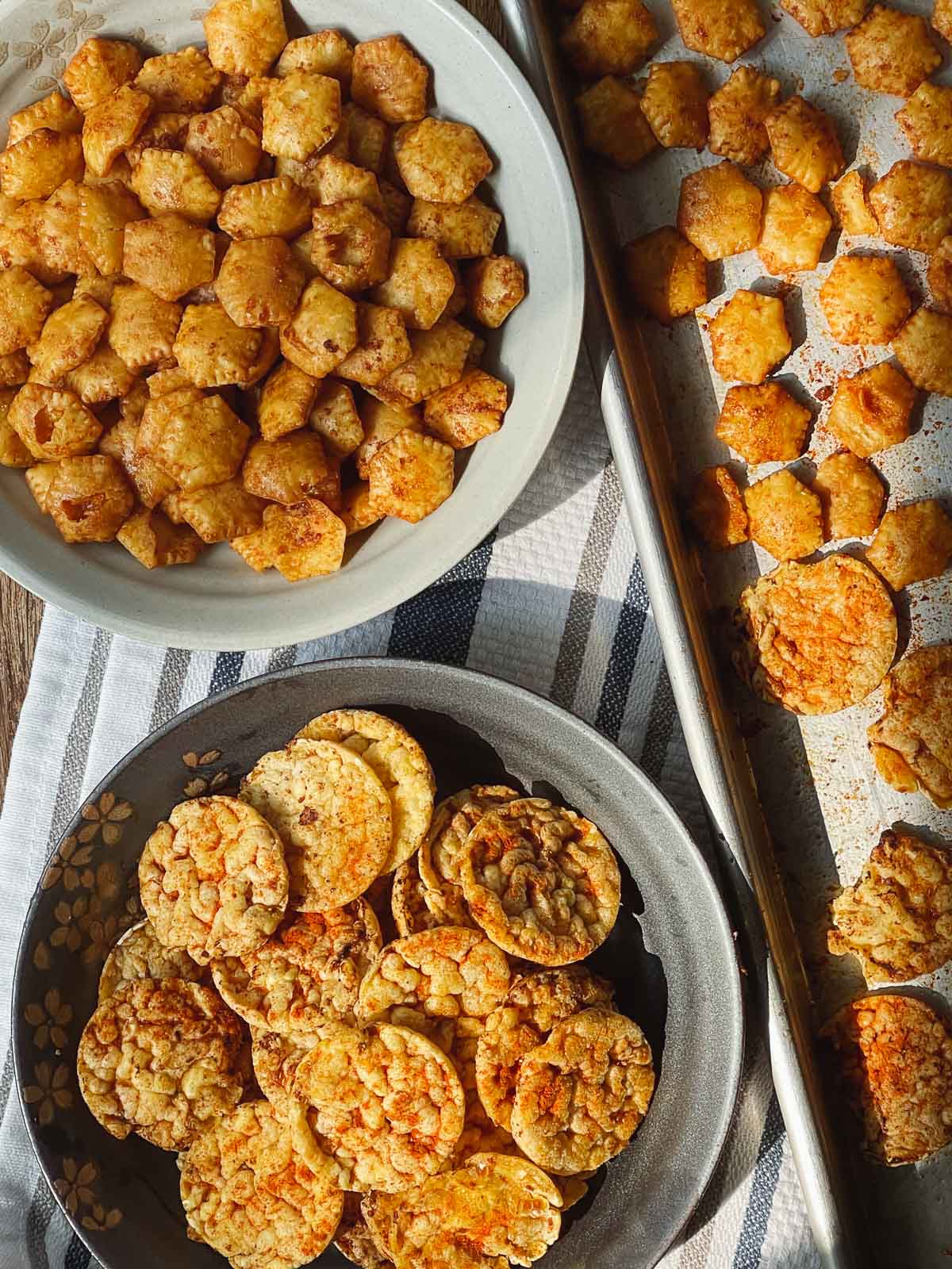 Sriracha popcorn cakes in a gray bowl and Sriracha oyster crackers in a light gray bowl on top of a striped kitchen towel and a baking tray with more crackers on the side.