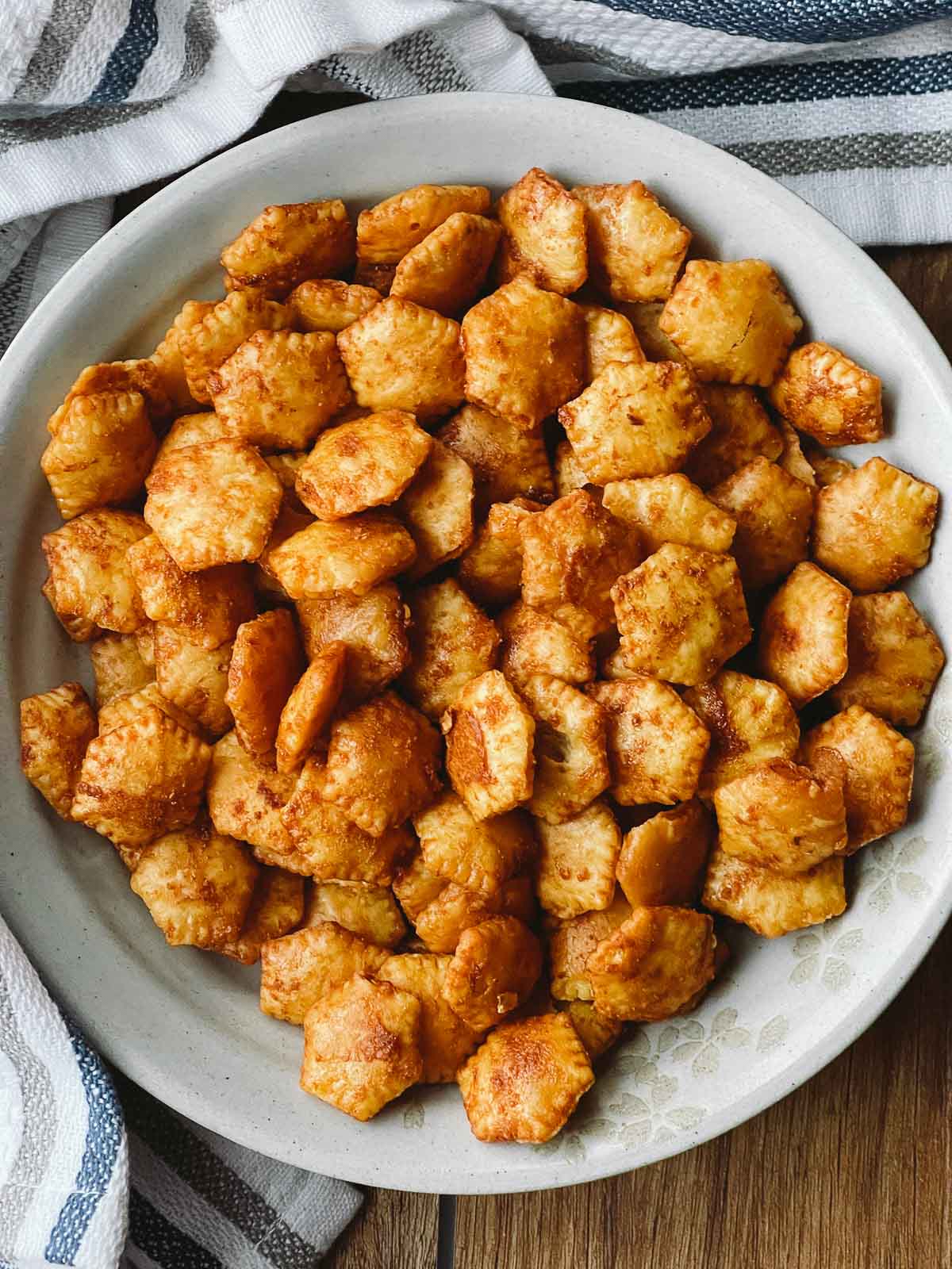 A white bowl filled with sriracha oyster crackers on a wooden board with a striped kitchen towel on the side.