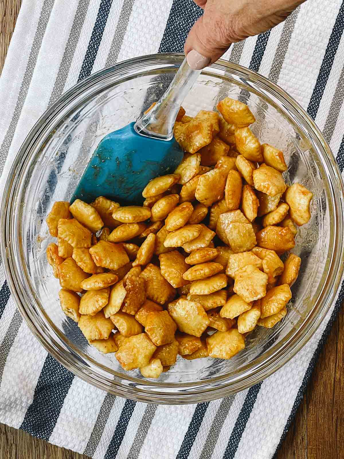 A blue silicone spatula stirring sriracha oyster crackers in a glass bowl on top of a striped kitchen towel.