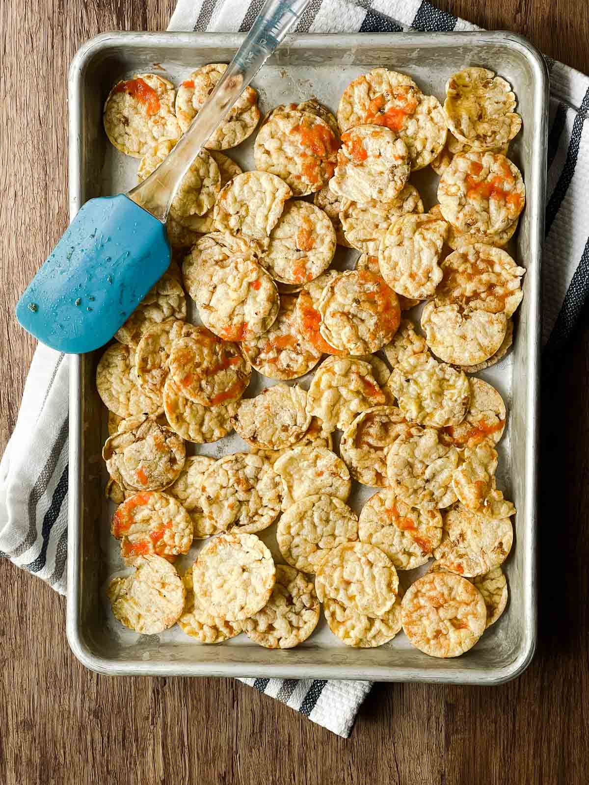 Sriracha mini popcorn cakes on a baking tray ready for the oven with a blue silicone spatula on the side on top of a striped kitchen towel.