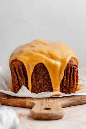 A loaf of pumpkin gingerbread topped with a caramel glaze on top of a wooden cutting board.