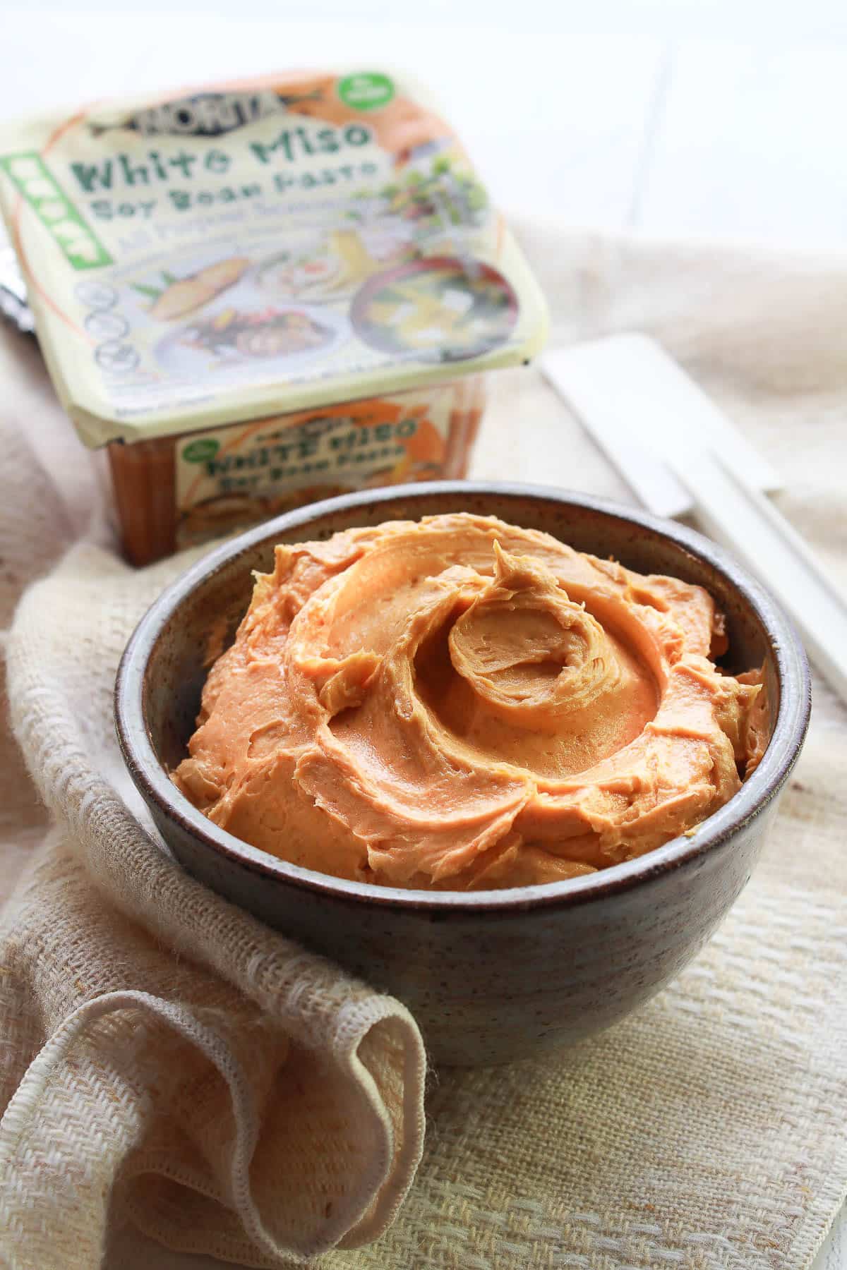 Miso butter in a brown bowl with miso paste in a package on the side, on top of a textured napkin