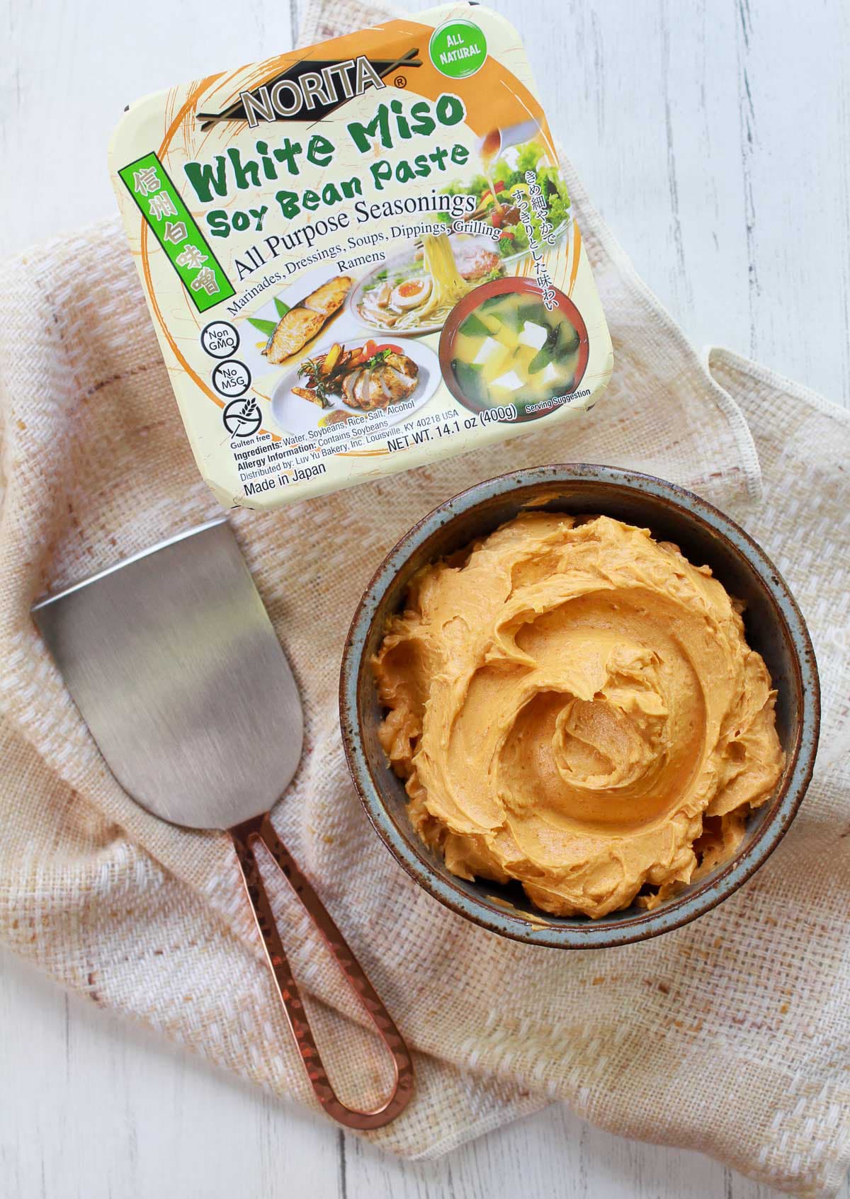 Miso butter in a brown bowl with miso paste in a package and a spreader on the side, on top of a textured napkin