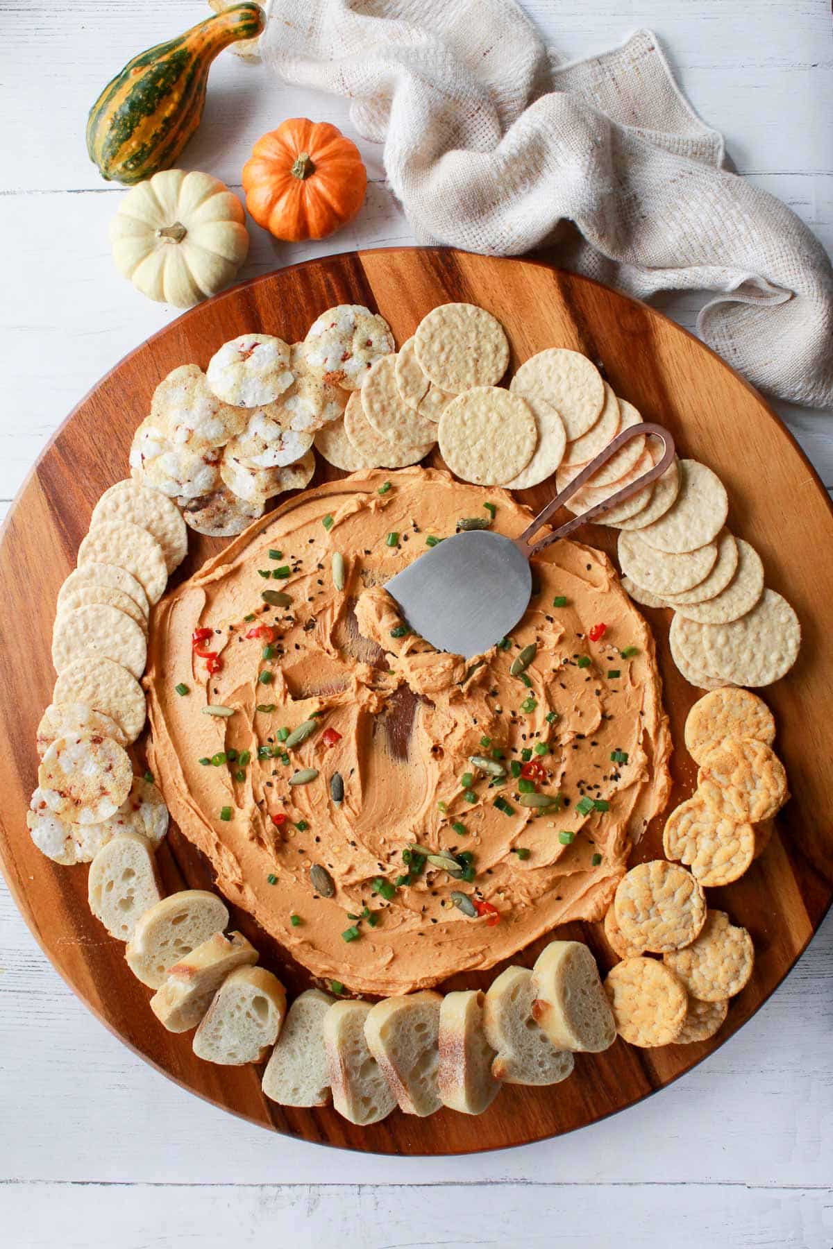 a miso butter board on a round wooden board surrounded by crackers and bread with baby pumpkins and a napkin on the side