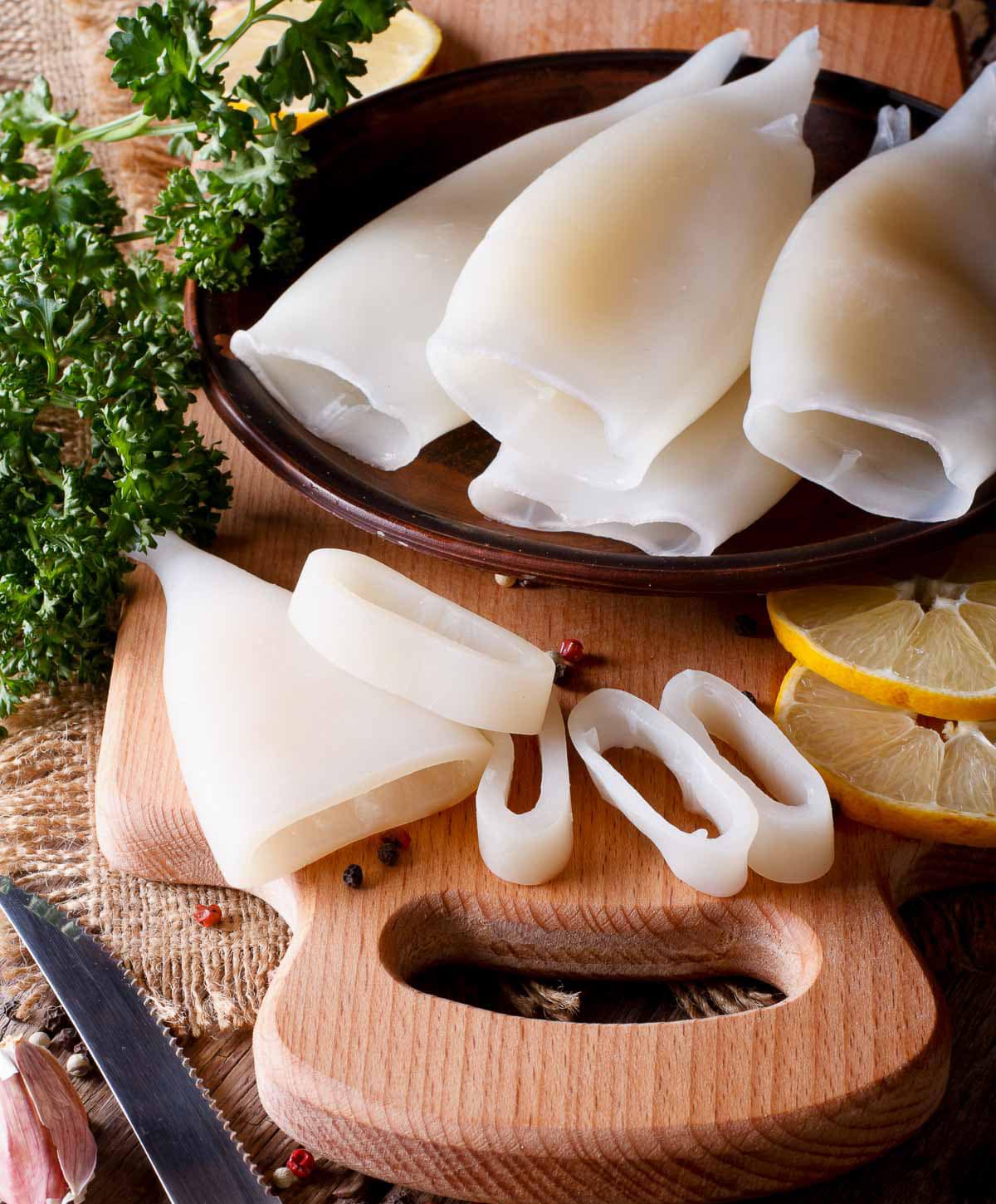 Whole raw squid and sliced squid rings on top of a wooden board with a knife and lemon slices on the side.