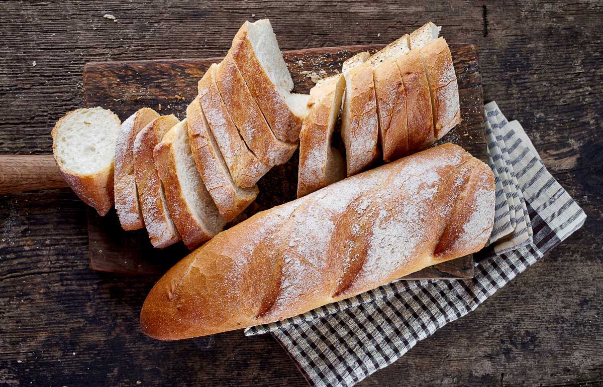 a loaf and slices of rustic, crusty bread on a wooden board with a checkered napkin on the side