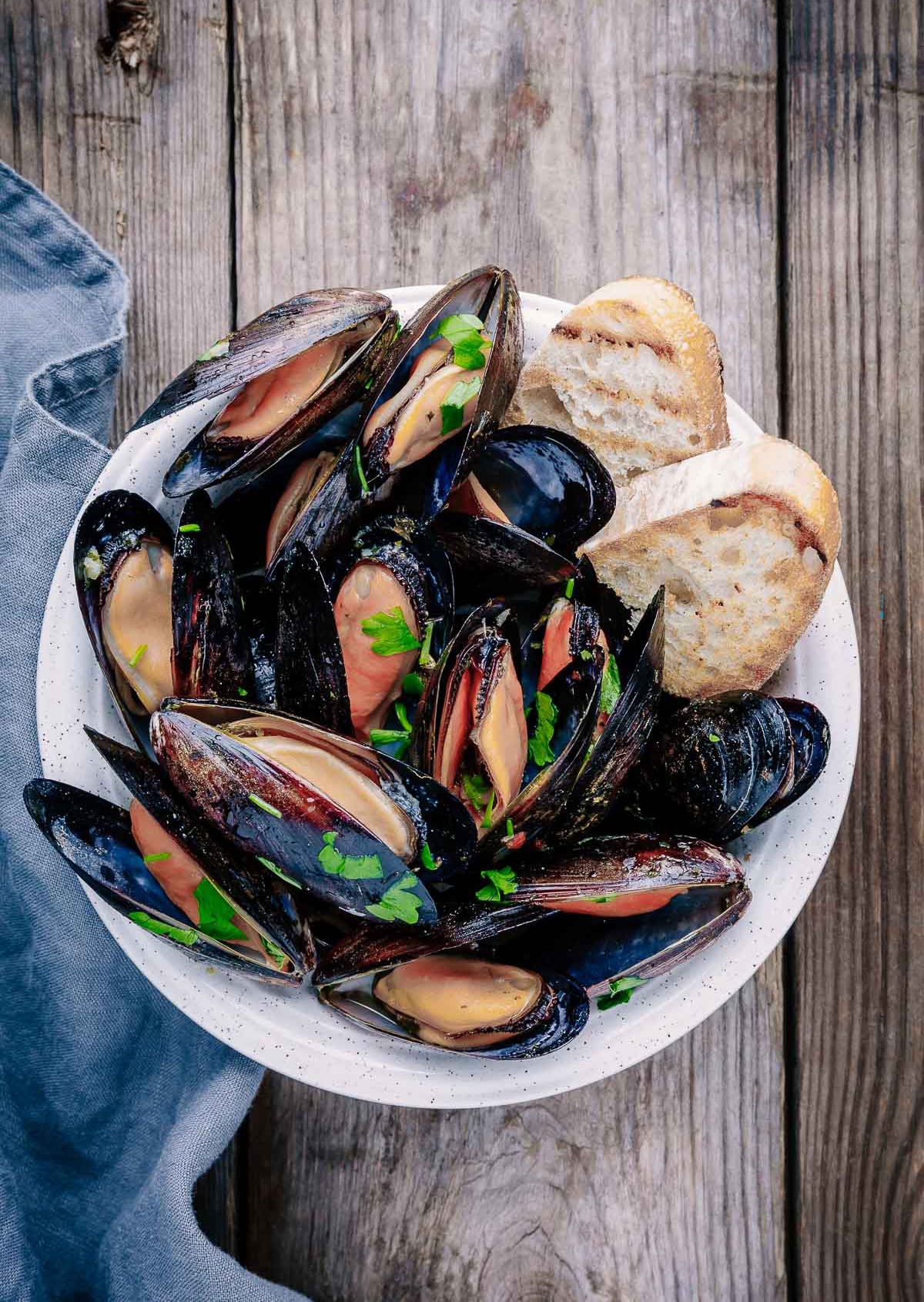 Thai curry steamed mussels presented in a white bowl with crusty bread on the side on top of a wooden board with a blue napkin