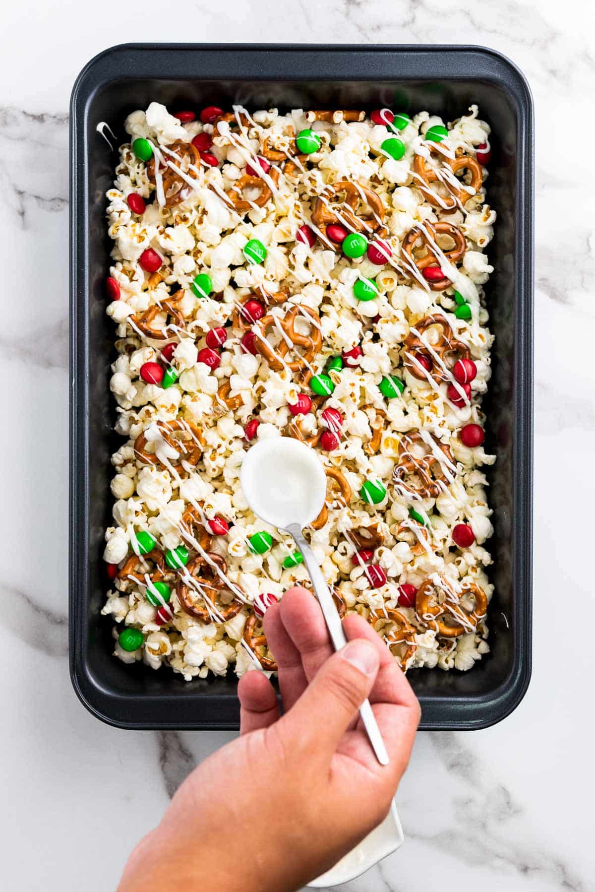Drizzling white chocolate on top of Christmas popcorn Santa munch in a black baking tray on top of a marble surface.