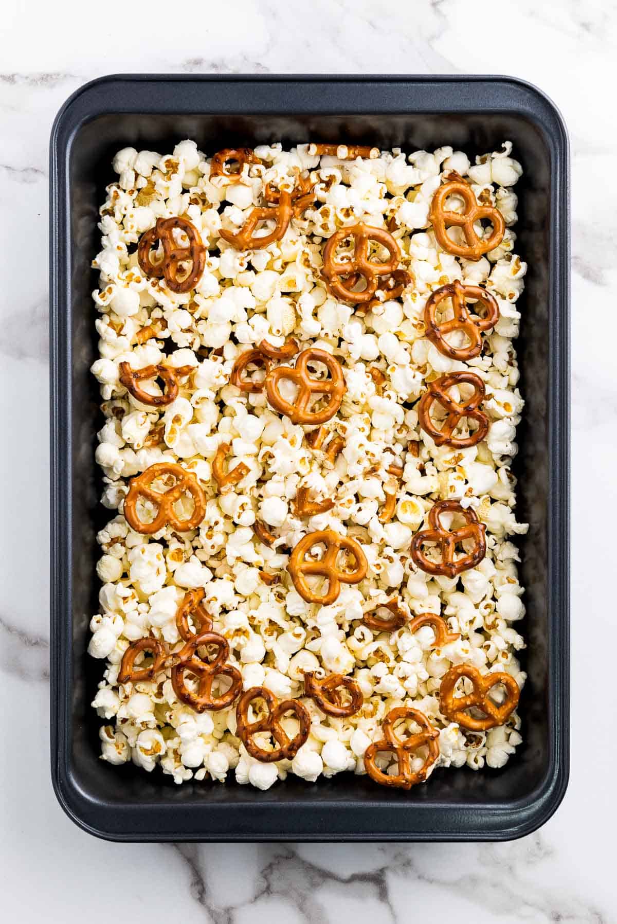 Popcorn and mini pretzels in a black baking tray on top of a marble surface.