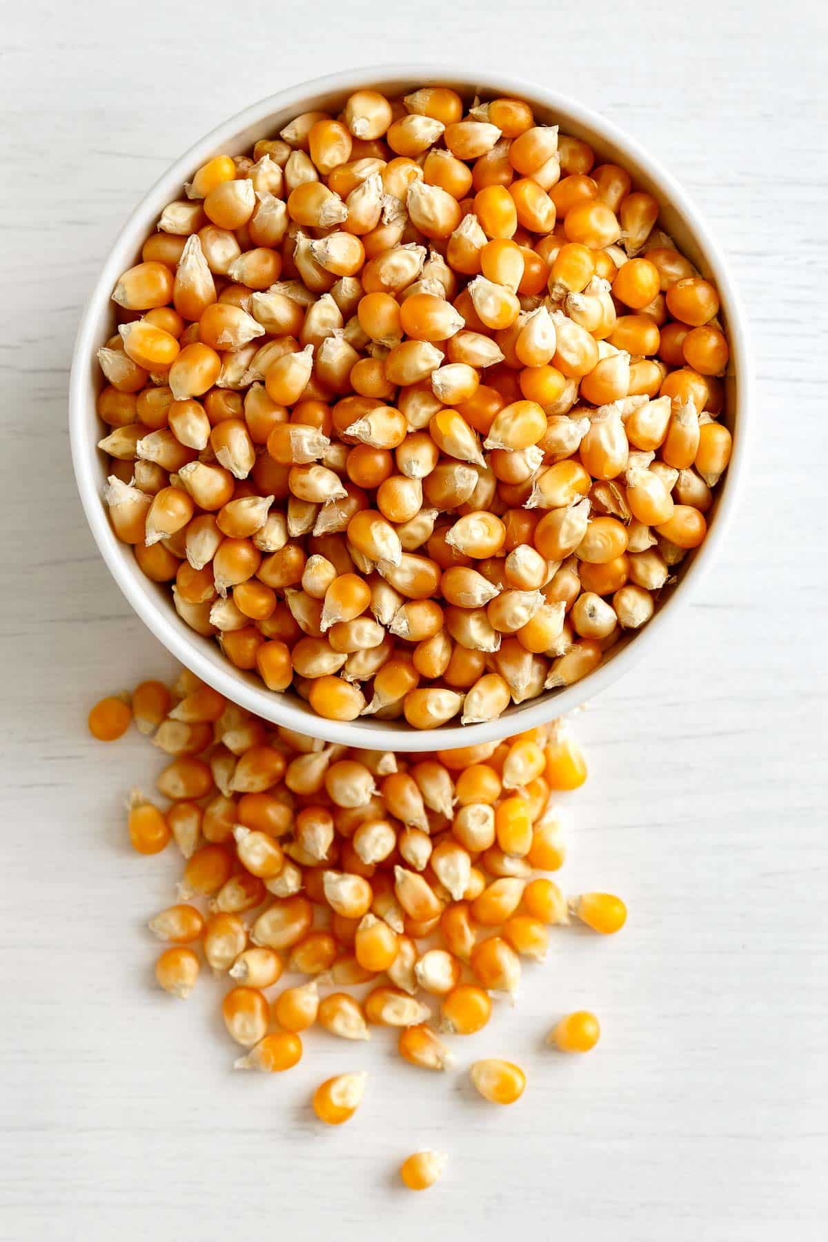 Unpopped popcorn in white bowl on painted white wood and spilled popcorn on the side.