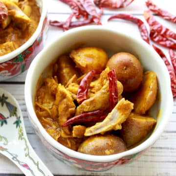 Two bowls filled with Chinese curry chicken and potatoes with whole red chili peppers on top all placed on a white wooden board with a white spoon on the side.