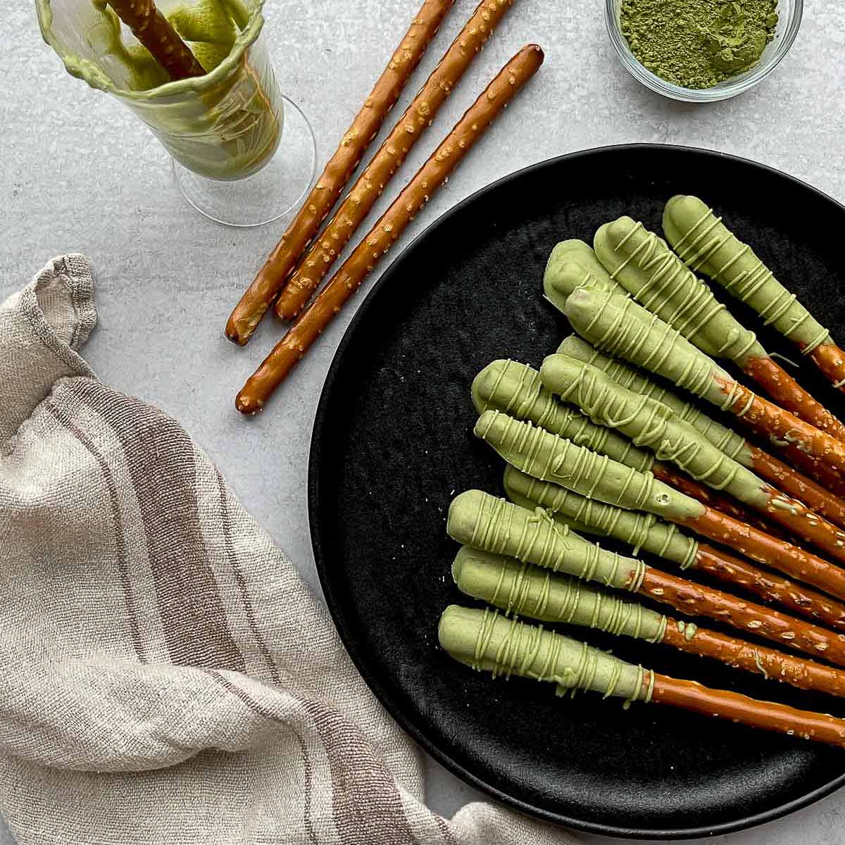 Matcha chocolate dipped pretzel rods on top of a round black plate with plain pretzel rods on the side, and a small bowl of matcha powder and a glass with a dipped pretzel rod, and a linen napkin on the side.