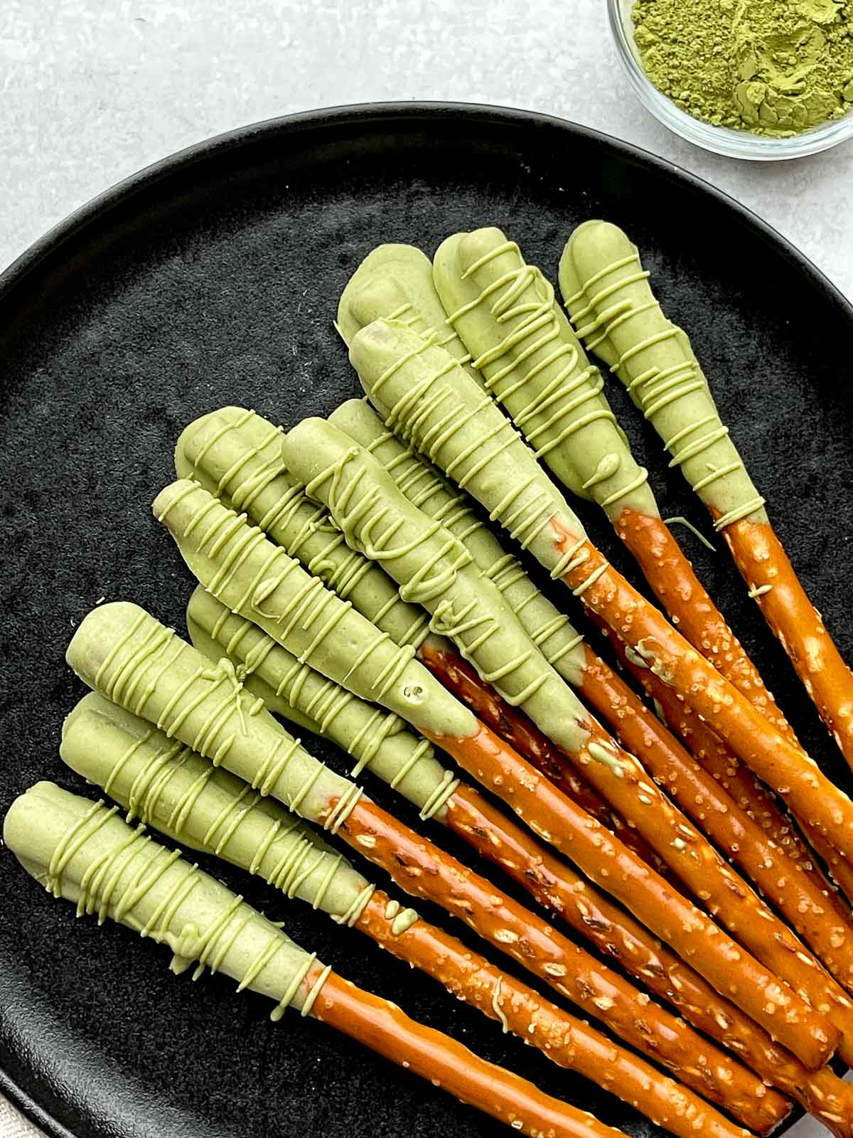Matcha chocolate dipped pretzel rods on top of a round black plate with plain pretzel rods on the side, and a small bowl of matcha powder and a linen napkin on the side.