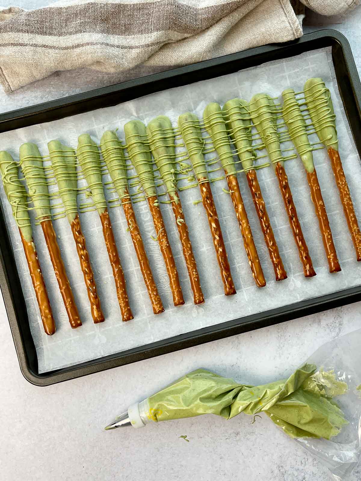 Matcha chocolate dipped pretzel rods lined up on top of parchment paper on a baking tray, with a frosting bag and linen napkin on the side.
