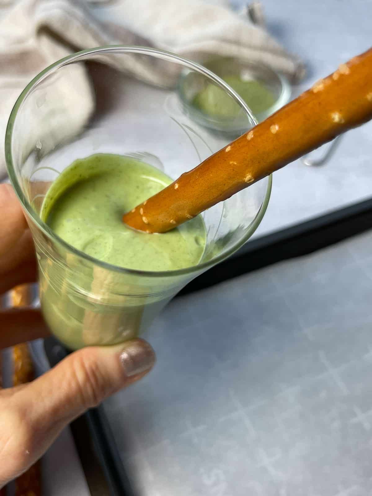 A pretzel rod being dipped into a tall glass with matcha chocolate above a baking tray lined with parchment paper.