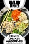 A large black bowl filled with Chinese chicken salad featuring shredded chicken, shredded carrots, broccoli slaw, scallions, water chestnuts and chow mein noodles, with a pair of brown chopsticks on top and a linen napkin on the side.
