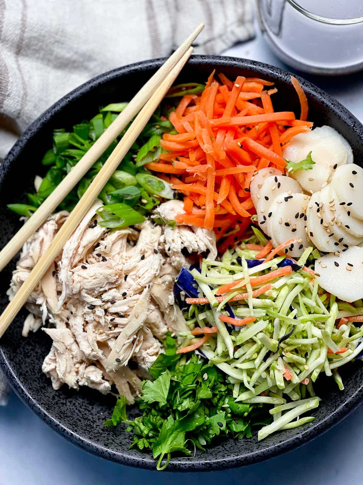 A large black bowl filled with Chinese chicken salad featuring shredded chicken, shredded carrots, broccoli slaw, scallions, water chestnuts and chow mein noodles, with a pair of brown chopsticks on top and a linen napkin on the side.