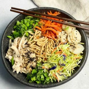 A large black bowl filled with Chinese chicken salad featuring shredded chicken, shredded carrots, broccoli slaw, scallions, water chestnuts and chow mein noodles, with a pair of brown chopsticks on top and a linen napkin on the side.