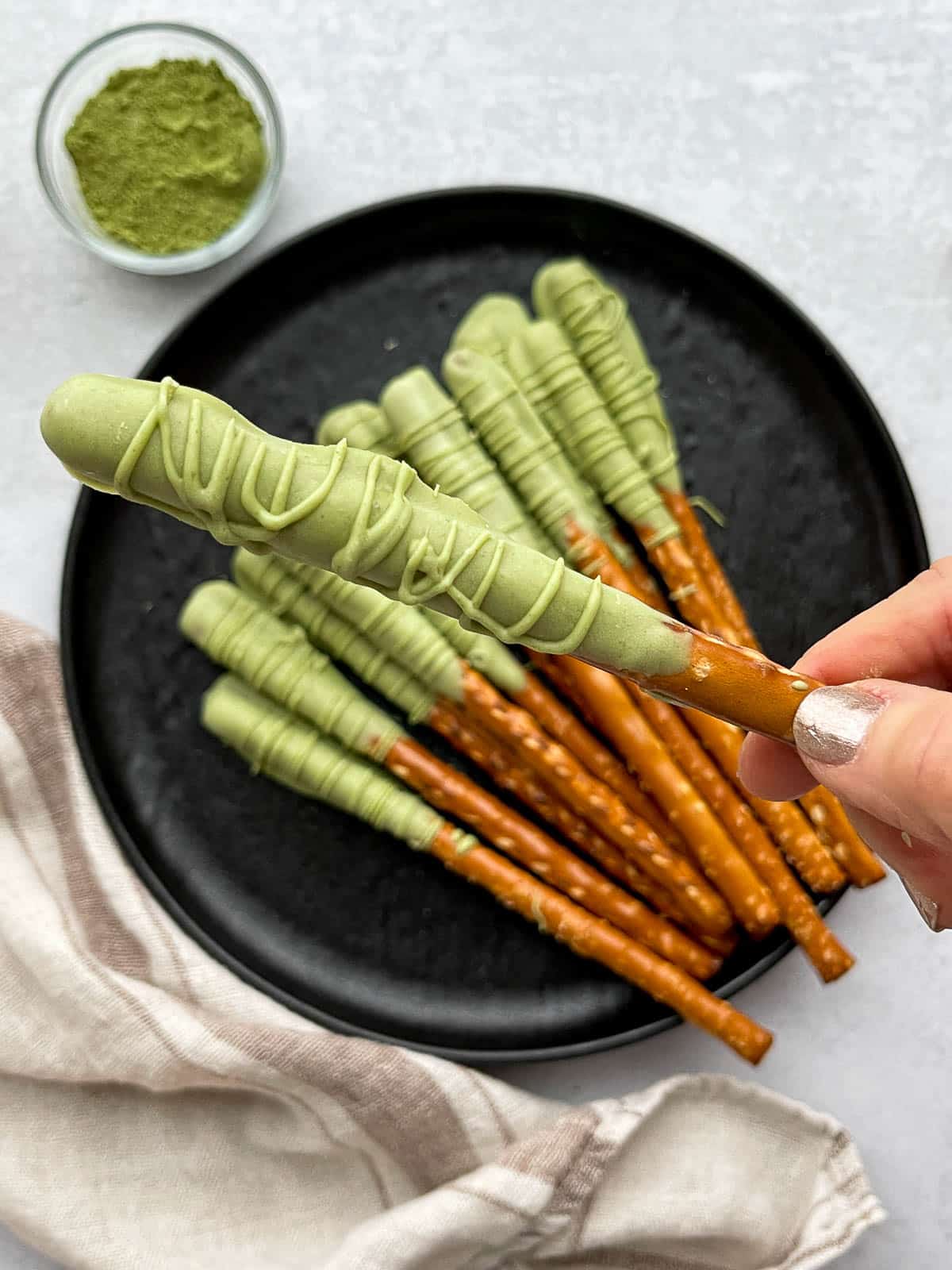 A hand holding up a chocolate matcha dipped pretzel rod above a round black plate with other dipped pretzel sticks and a small bowl with matcha powder on the side with a linen napkin.