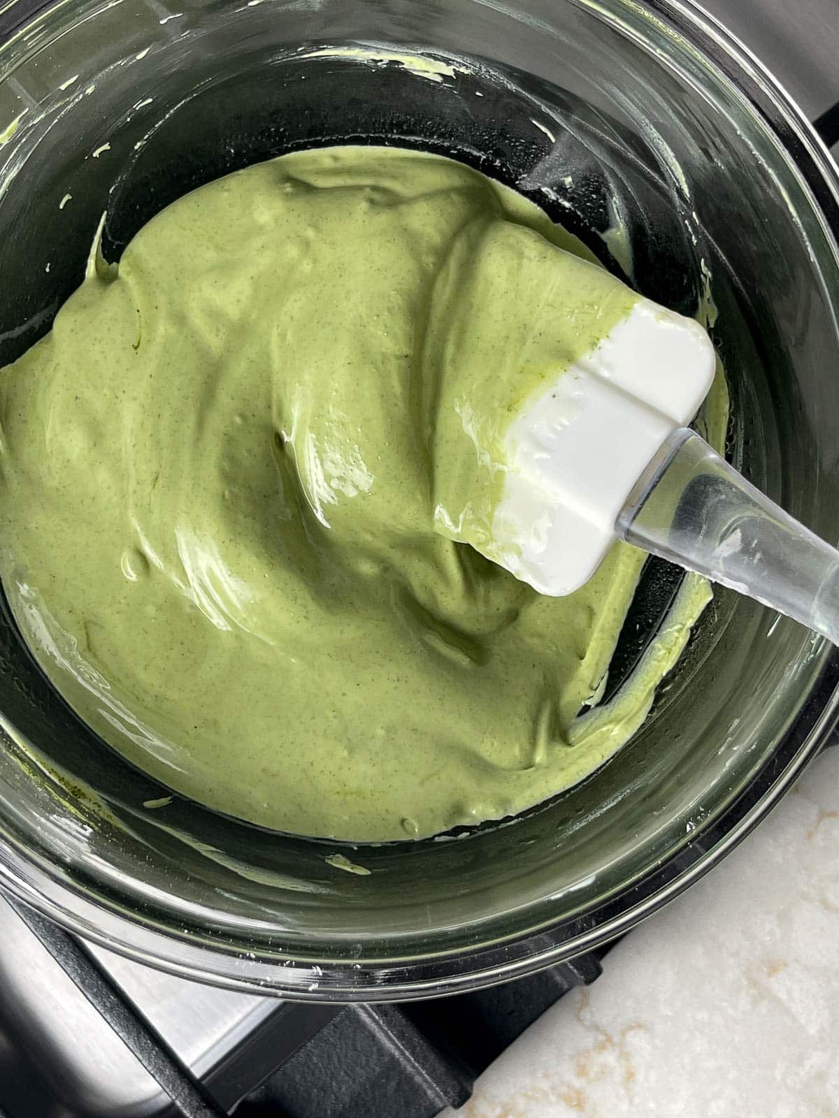 A white rubber spatula stirring melted matcha chocolate in a glass bowl on a stove top.