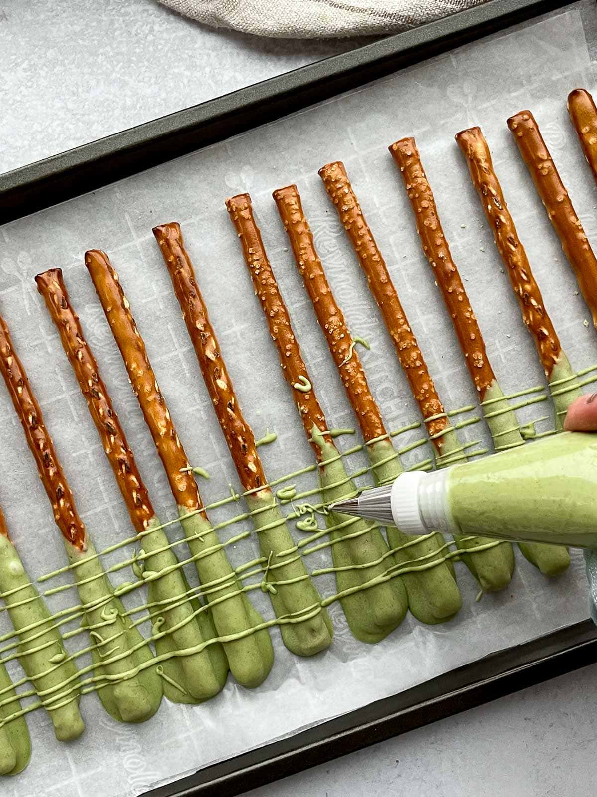 A piping bag filled with matcha chocolate piping over a row of pretzel rods on top of parchment paper on a black baking tray.