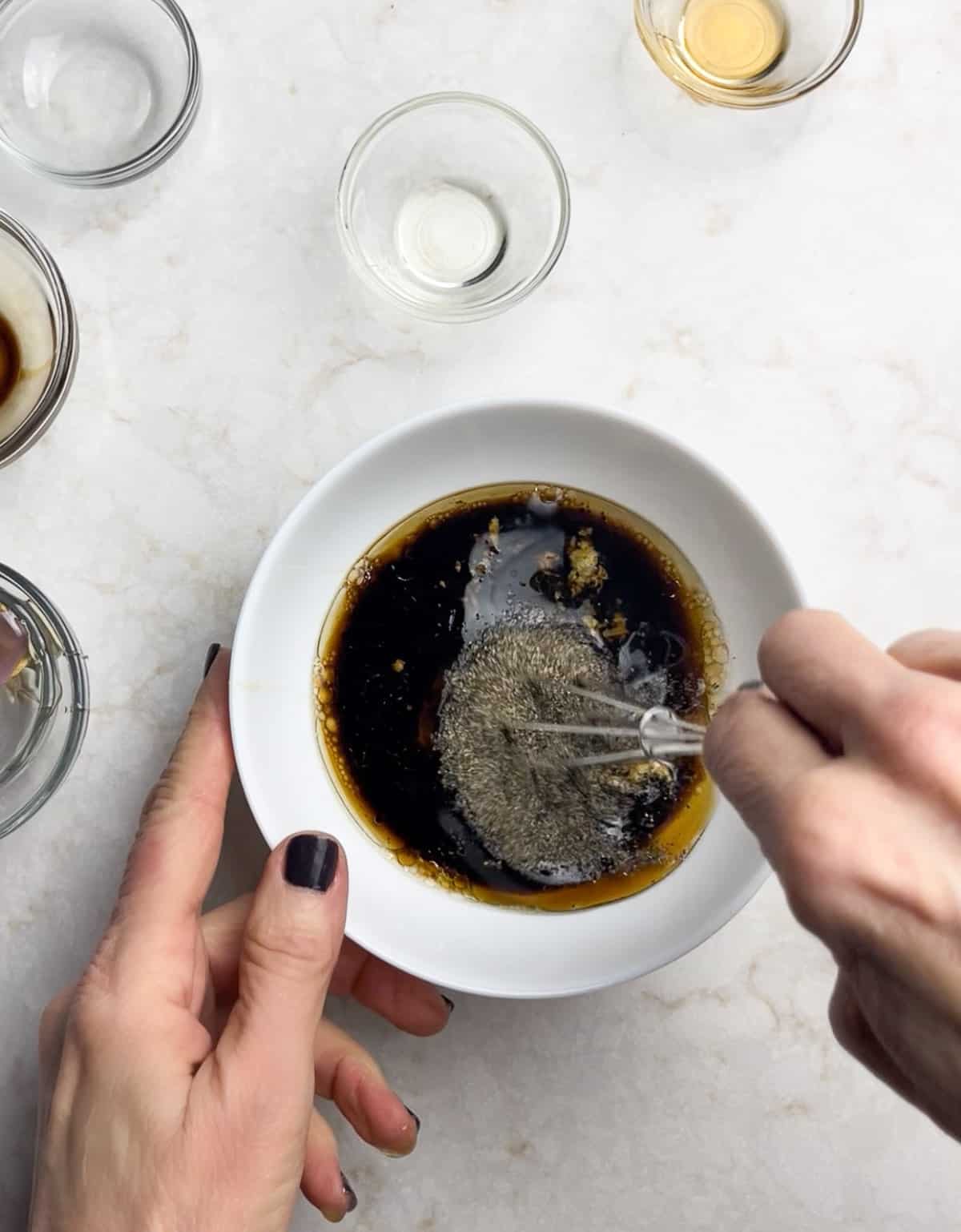 A woman's hands whisking the Asian salad dressing for Chinese chicken salad in a small white bowl, with other glass bowls surrounding.