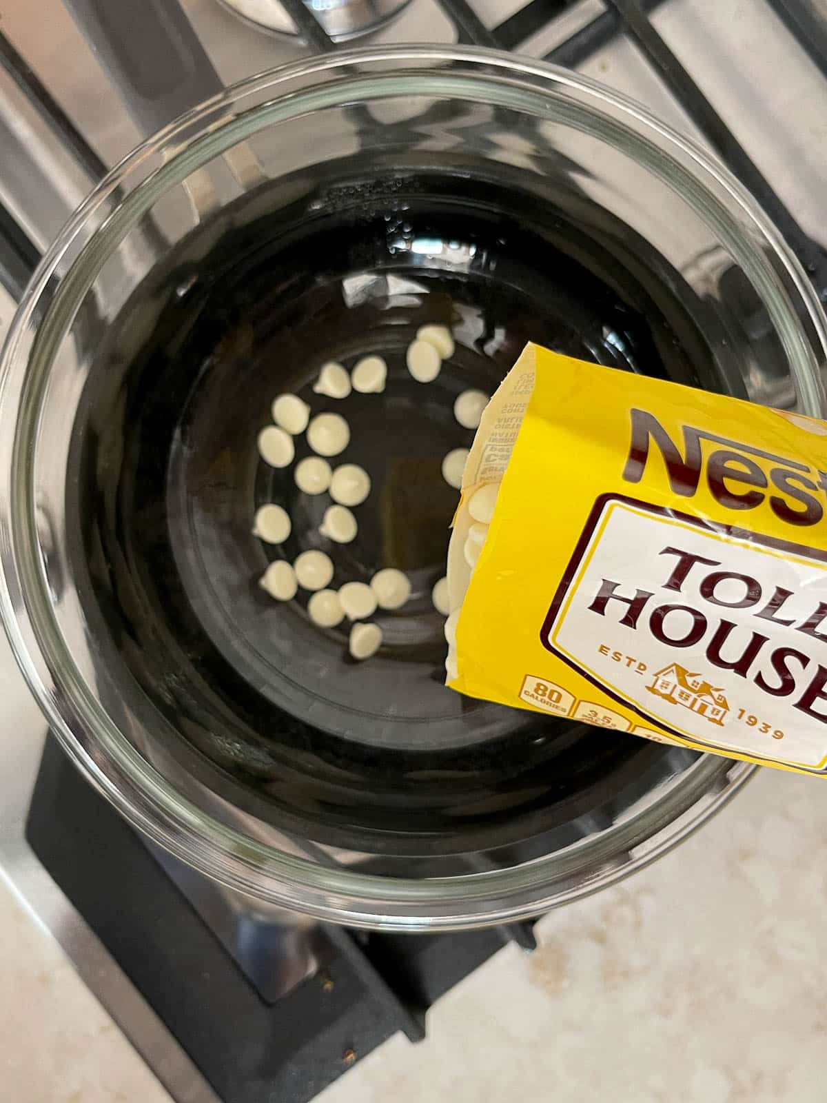 Nestle white chocolate morsels being poured into a large glass bowl over a stove top.