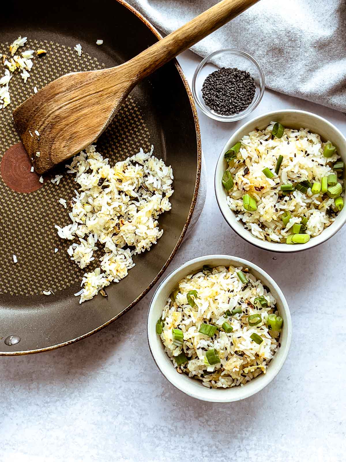 Crispy rice with ginger and scallions in two small bowls with chopsticks on top and a pan with rice with a wooden spatula, and black sesame seeds in a small bowl on the side.