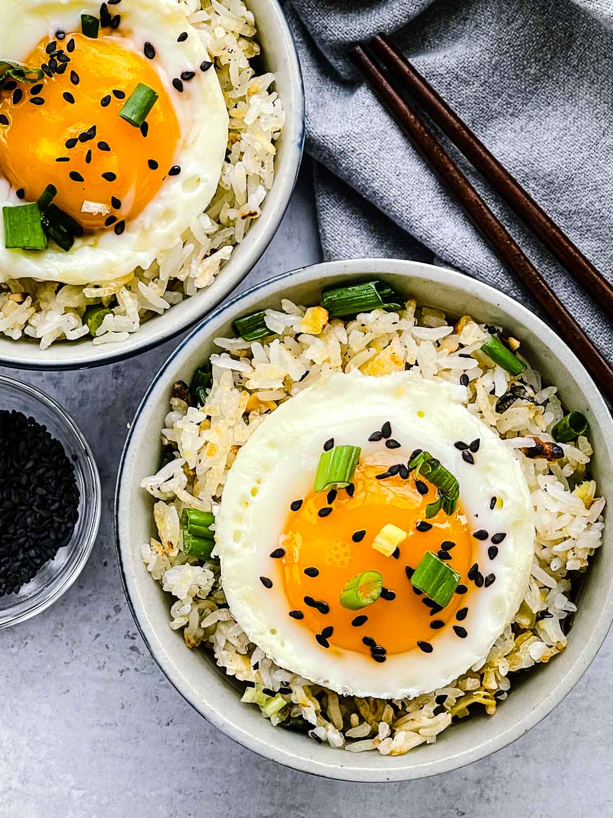 Crispy rice with ginger and scallions topped with a runny fried egg in two small bowls with a pair of brown chopsticks and black sesame seeds in a bowl on the side.
