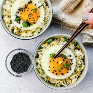 Crispy rice with ginger and scallions topped with a runny fried egg in two small bowls with a pair of brown chopsticks, a linen napkin, and black sesame seeds in a bowl on the side.
