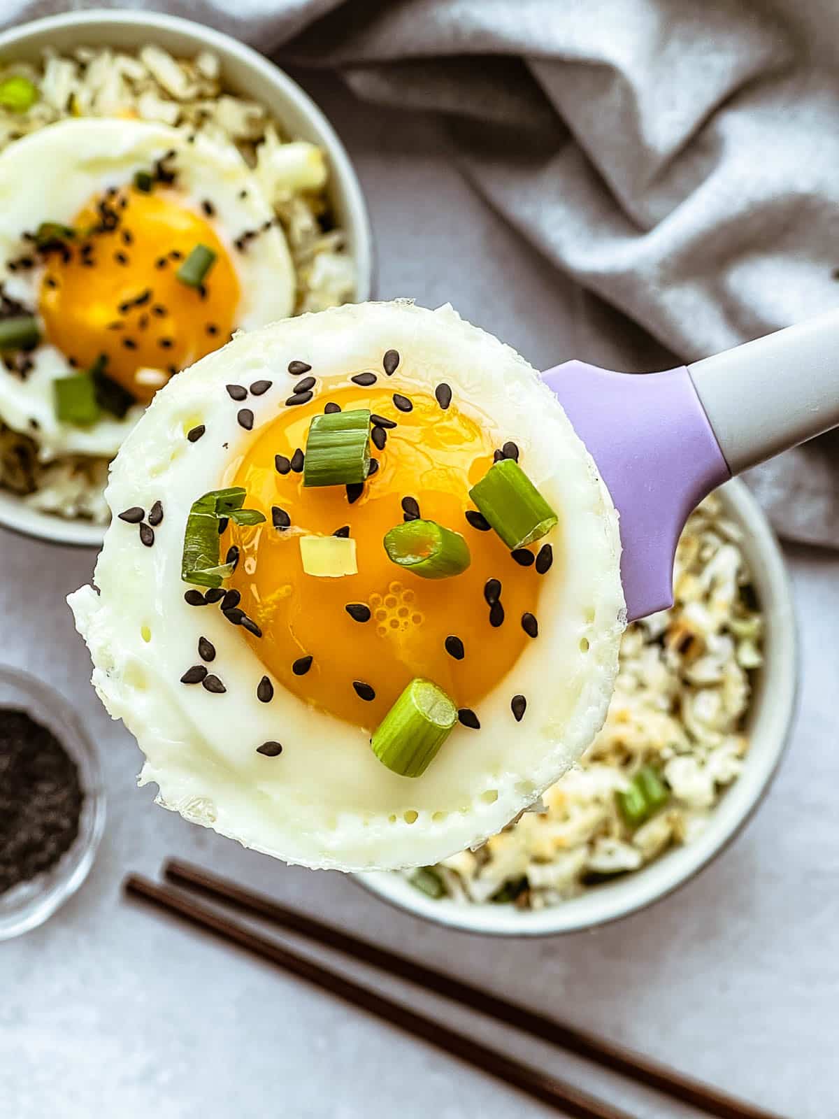 A close-up image of a runny fried egg above two bowls of crispy rice with ginger and scallions and chopsticks on the side.