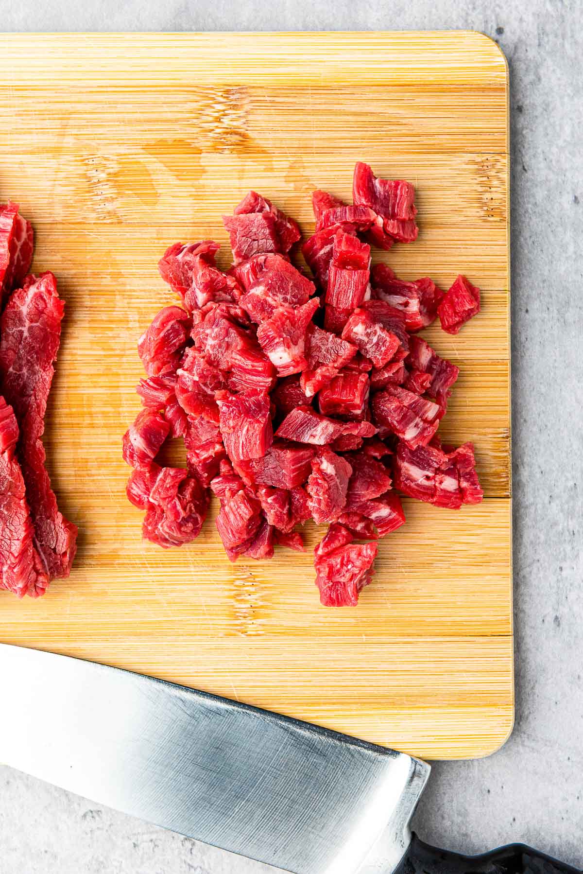Cubed flank steak on top of a wooden cutting board with a large knife on the side.
