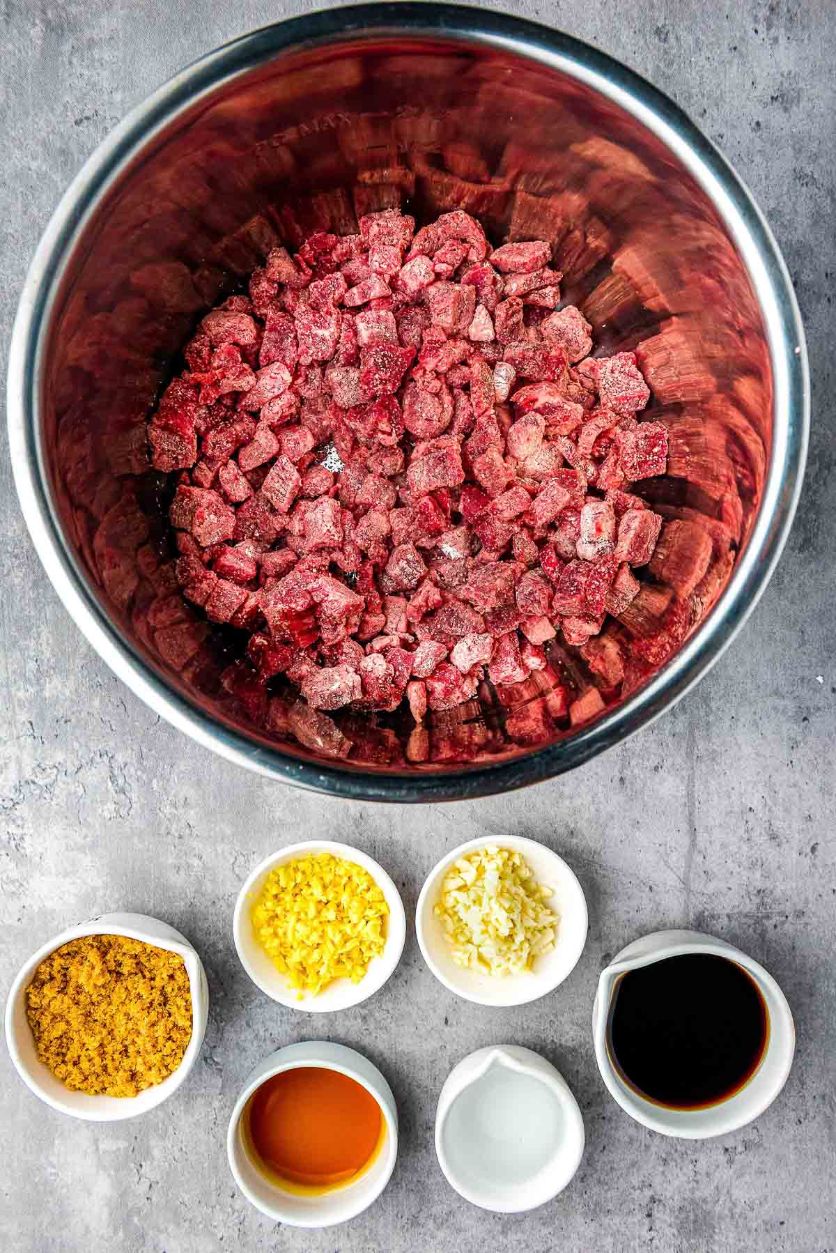 Cubed flank steak marinating in a silver bowl with marinade ingredients in small bowls on the side.