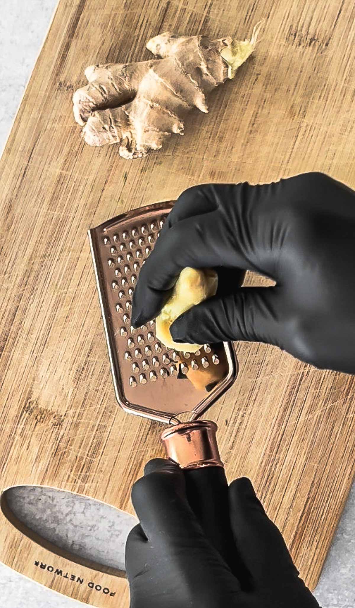 Hands with black gloves grating fresh ginger on top of a wooden cutting board, with a whole ginger bulb on the side.