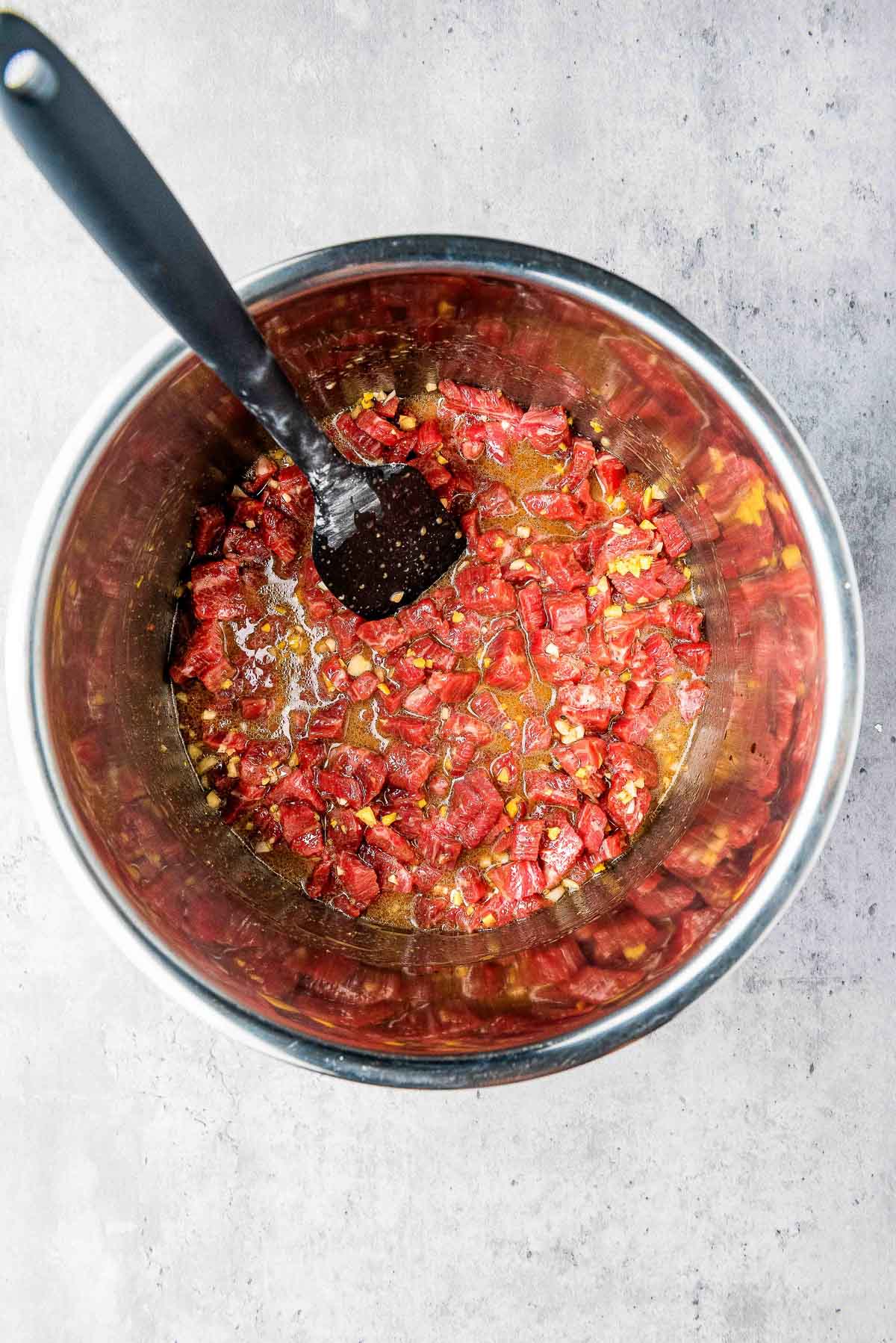 Marinated cubed flank steak in a silver bowl with a black spoon, on top of a gray surface.