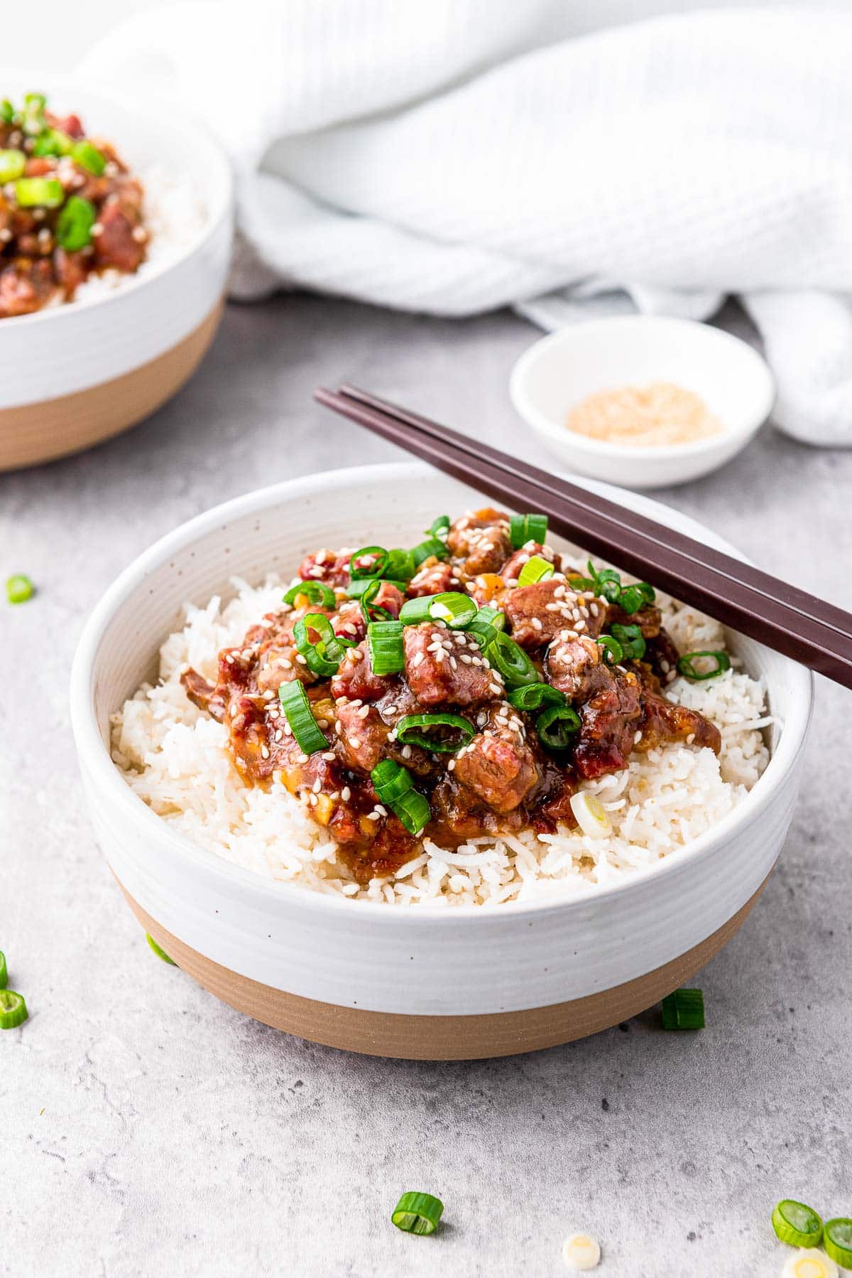 A pair of chopsticks on top of a white bowl filled with Slow Cooker Mongolian Beef on white rice, with a bowl of Mongolian beef in the background on top of a gray surface.