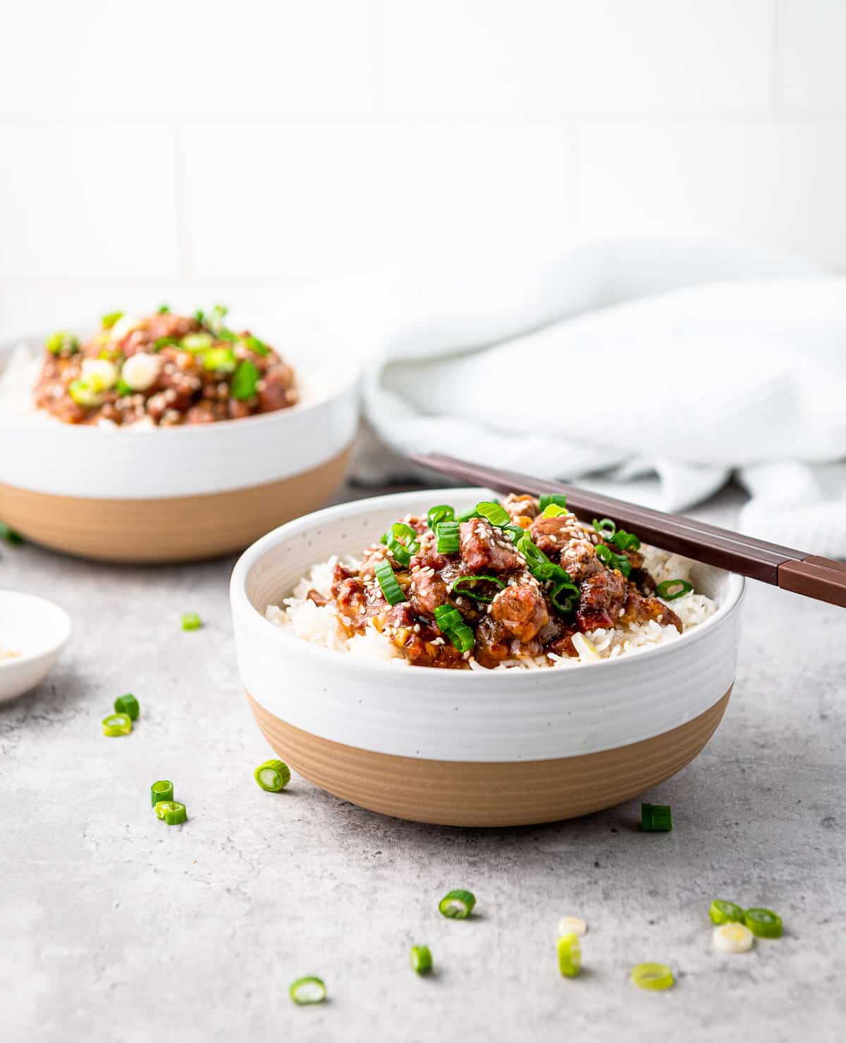 A pair of chopsticks on top of a white bowl filled with Slow Cooker Mongolian Beef on white rice, with a bowl of Mongolian beef in the background on top of a gray surface.