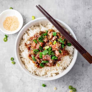 A pair of chopsticks on top of a white bowl filled with Slow Cooker Mongolian Beef on white rice, with a bowl of Mongolian beef in the background on top of a gray surface.