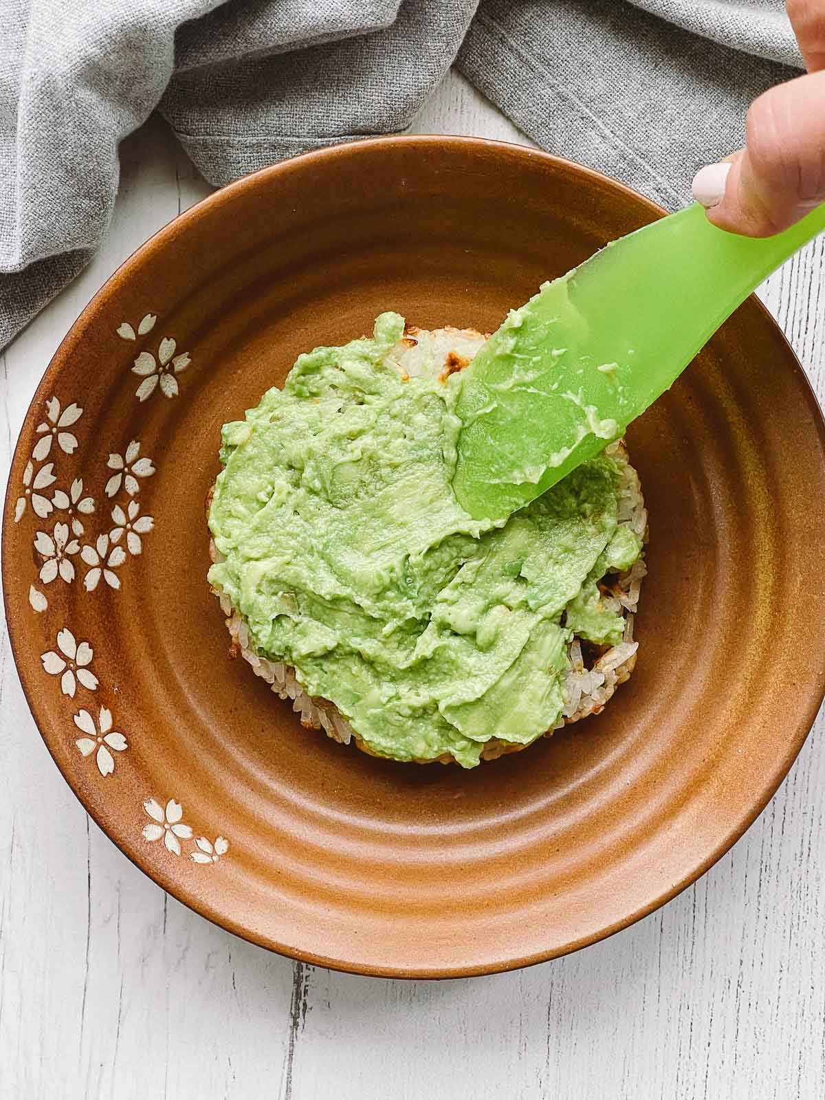 A green spatula spreading mashed avocado on top of a rice waffle on a brown plate with a linen napkin on the side.